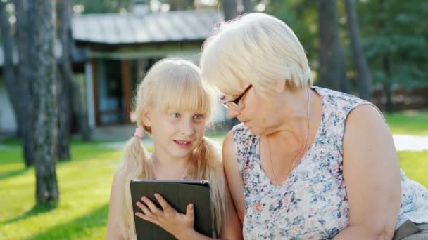 Oma en kleindochter rusten in de achtertuin van het huis, ze kijken naar de tablet samen, communiceren. — Stockvideo