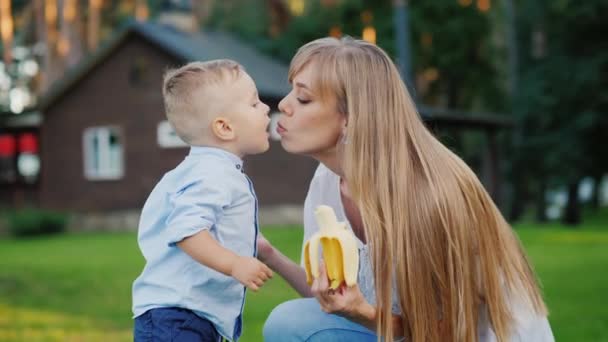 El niño besa a su madre. Descansan en el patio de su casa, el niño se comió un plátano — Vídeo de stock