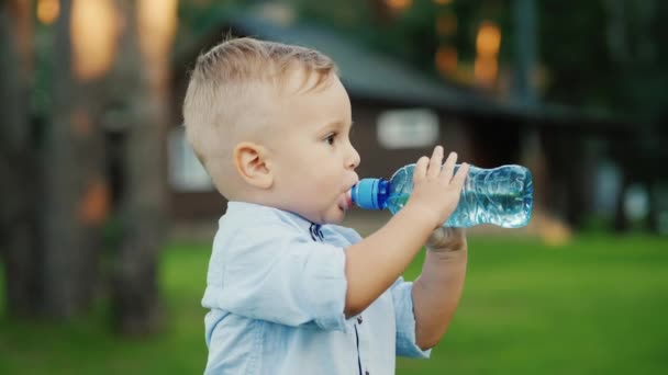 Il bambino beve acqua dalla bottiglia da un anno. In piedi nel cortile di casa tua — Video Stock