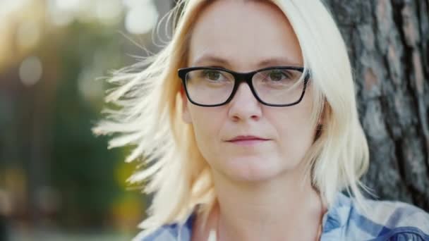 Retrato de una joven pensativa en gafas. Él mira a lo lejos, el viento juega con su pelo. Se sienta en un parque cerca de un árbol. Vídeo en cámara lenta — Vídeos de Stock