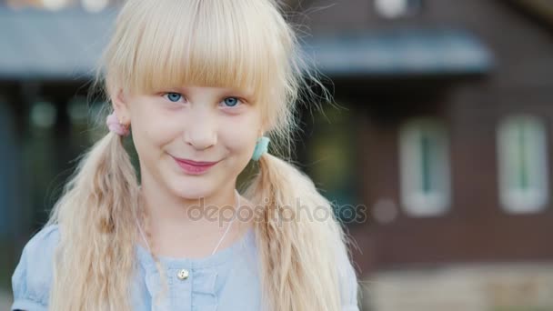 Retrato de uma menina loira de 6 anos, sorrindo para a câmera. Fica no fundo da casa dele. Vídeo com profundidade de campo rasa — Vídeo de Stock