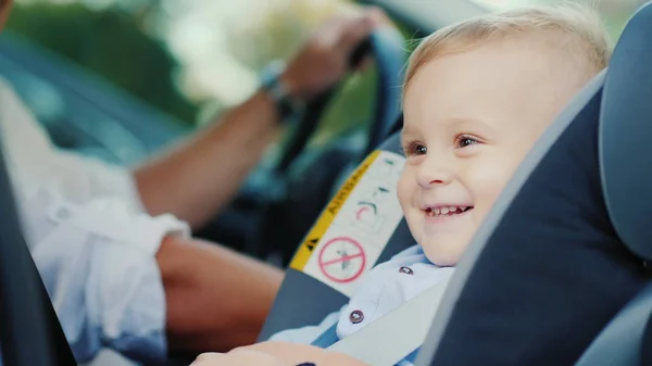 Un niño pequeño está sentado en un asiento de coche cerca de su padre, sonriendo felizmente. Concepto - seguridad y cuidado — Foto de Stock