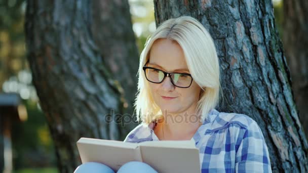 En lyshåret ung kvinde med briller læser en bog i parken. Sidder nær et træ, smukt lys før solnedgang – Stock-video