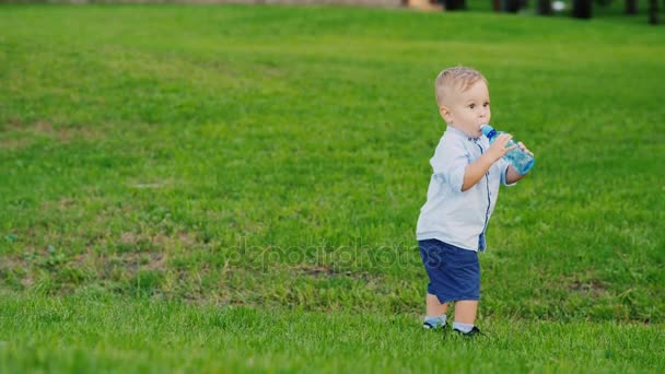 Funny kid 1 year is drinking water from a bottle. It stands on a green lawn, coolly staggers. Fun video with children — Stock Video