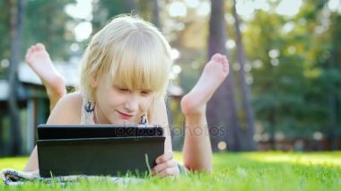 6 yıl eski, bir tablet üzerinde oynayan bir kız evinin yakınında yeşil çim üzerinde yatıyor