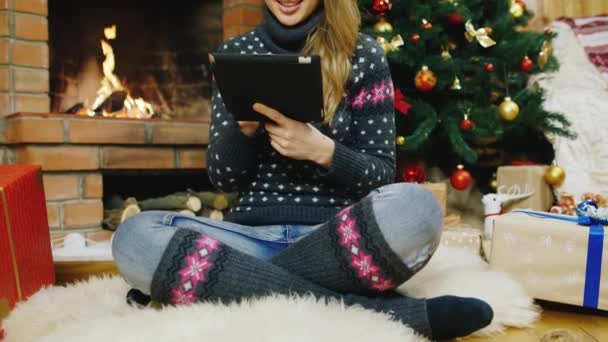 Ein gemütliches Haus mit Kamin und Weihnachtsbaum. Neben dem Tablet steht eine hübsche Frau. Konzept - Online-Geschenkbestellung — Stockvideo