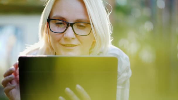 Uma jovem mulher de óculos jaz em um gramado no parque, desfruta de um tablet. Tecnologia na natureza — Vídeo de Stock