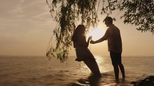 Boas férias juntos. Um jovem casal está montando em um balanço em uma praia tropical. Pôr do sol, há uma chuva quente — Vídeo de Stock