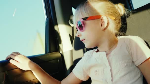 Uma menina de 6 anos de idade com óculos rosa protetores contra o sol está andando no banco de trás do carro. Fixa-se com um cinto de segurança — Vídeo de Stock