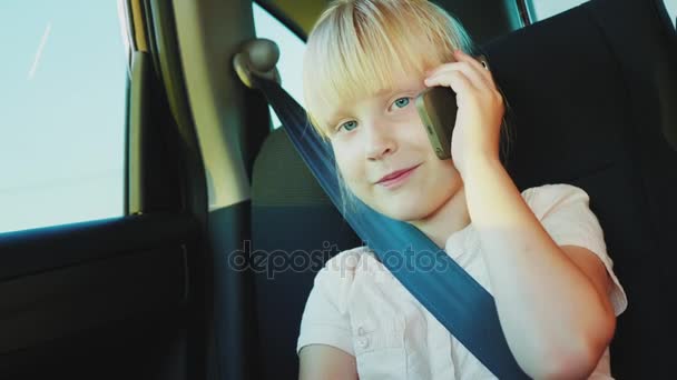 Забавная 6-летняя девочка говорит по телефону, едет на заднем сиденье машины. Пристегивается ремнем безопасности — стоковое видео