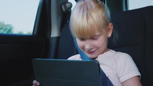 Diversão na estrada. A menina usa um tablet, cavalga no banco de trás do carro — Vídeo de Stock