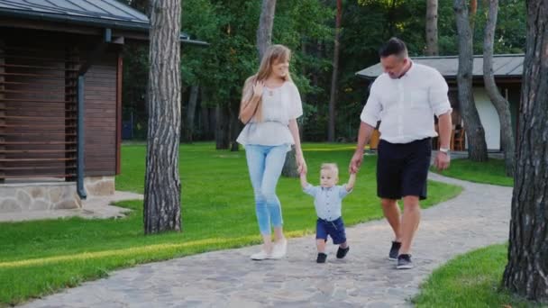 En ung familj med en liten son promenader runt deras hus. De hålla barnet i handen, promenera längs vägen — Stockvideo