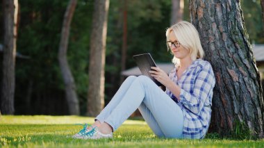 Genç bir kadın bir tablet kullanıyor. Evin arka bahçesinde bir ağacın altında çimlerin üzerine oturur