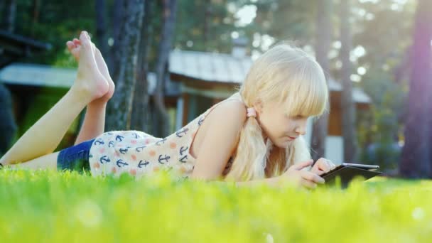 无忧无虑的金发女孩正在玩这款平板电脑。在房子附近的绿色草地上的谎言，太阳精美照亮她的头发 — 图库视频影像