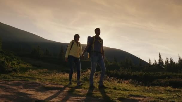 Fiatal pár turista kézenfogva, séta a lenyugvó nap sugaraiban egy hegyi ösvény — Stock videók