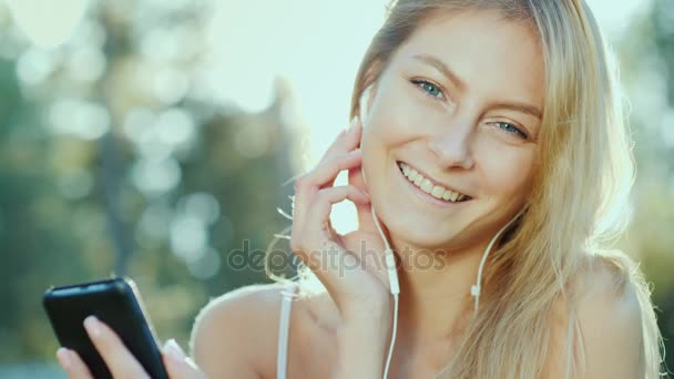 Mladá žena ve sluchátkách poslouchá hudbu, používá smartphone. Slunce krásně svítí, její vlasy — Stock video