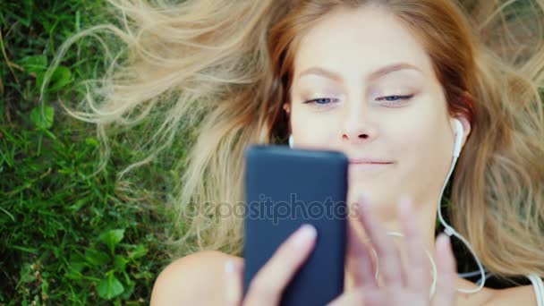 Aantrekkelijke jonge vrouw ligt op gras, geniet van de smartphone. Haar prachtig liggen op het gazon — Stockvideo