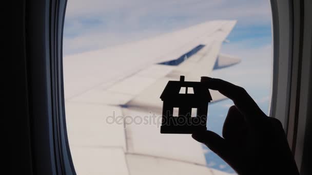 Eine weibliche Hand hält die Figur eines kleinen Hauses auf dem Hintergrund eines Flugzeugfensters. Flügel und der Himmel im Hintergrund — Stockvideo