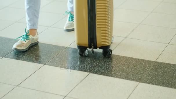 Een vrouwelijke reiziger met een tas op wielen is een wandeling langs de luchthaventerminal. In het frame zijn alleen de benen zichtbaar — Stockvideo