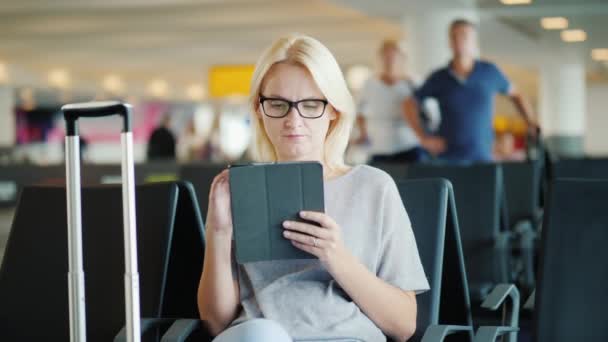 Uma jovem de óculos usa um tablet. Senta-se no terminal do aeroporto, ao lado de seu saco de viagem sobre rodas. À espera de um voo — Vídeo de Stock