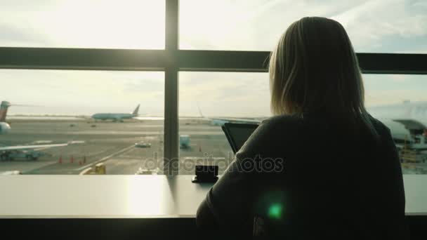 Une femme s'assoit à la fenêtre du terminal de l'aéroport à la table, profite d'une tablette. Toujours en ligne, confort de voyage — Video