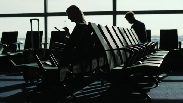 Le persone al terminal dell'aeroporto stanno aspettando il loro volo. Silhouette sullo sfondo della finestra, utilizzare il dispositivo — Video Stock