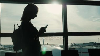 Her zaman online. Havaalanı terminalinde büyük bir pencerenin yanında akıllı telefon kullanan bir kadının silüeti.