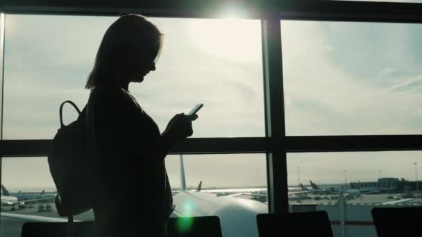 Always online. Silhouette of a woman using a smartphone in an airport terminal near a large window — Stock Video