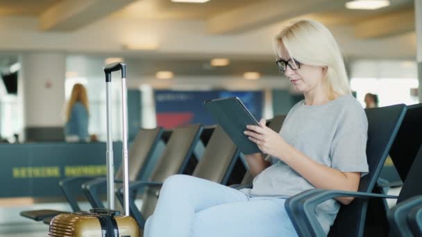 Bir kadın bir havaalanında terminal bir seyahat eder. Bir uçuş beklentisiyle oturur, bir tablet kullanır — Stok video