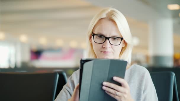 Retrato de uma mulher de óculos. Senta-se na sala de espera da estação de ônibus, usa um tablet — Vídeo de Stock