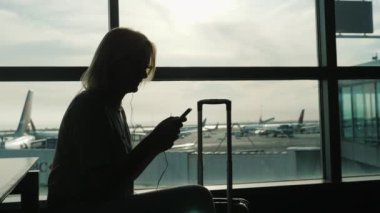 Büyük Havaalanı terminal penceresi oturan bir kadın silueti. Bir akıllı telefon kullanır. Uçuş için bekliyor