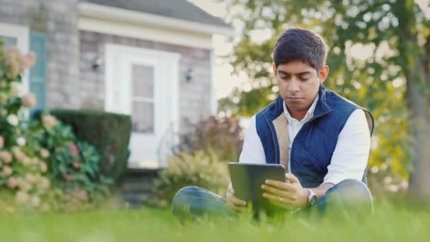Un Indien utilise une tablette dans la cour de sa maison. Assis sur la pelouse — Video