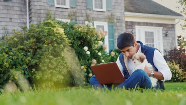 Un joven indio trabajando con un portátil en su patio. En sus manos tiene un cachorro. Por todas partes con la mascota — Vídeos de Stock