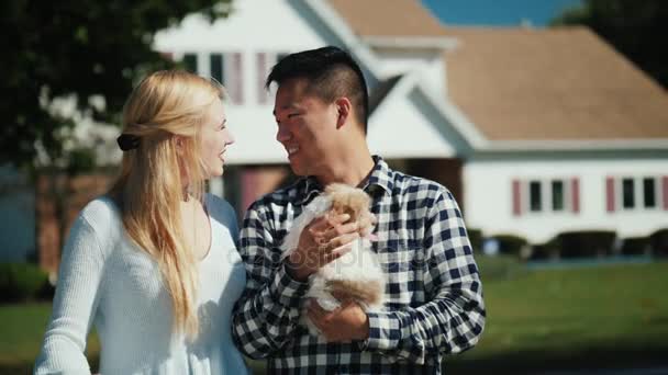 Gelukkige eigenaars van het nieuwe huis. Kaukasische vrouw met een Aziatische man op de achtergrond van een nieuw huis. Een man heeft een pup in zijn handen, en de sleutels van een vrouw — Stockvideo