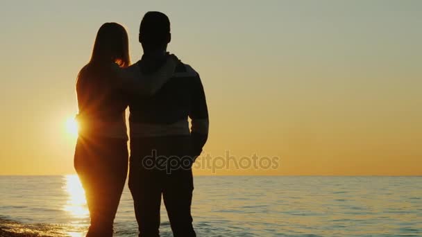 Silhuetas de um jovem casal abraçando e admirando o pôr do sol junto ao mar. Visão traseira — Vídeo de Stock