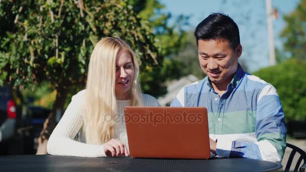 Los jóvenes caucásicos mujer y el hombre chino están utilizando el ordenador portátil juntos. Descansa en un café al aire libre. — Vídeo de stock