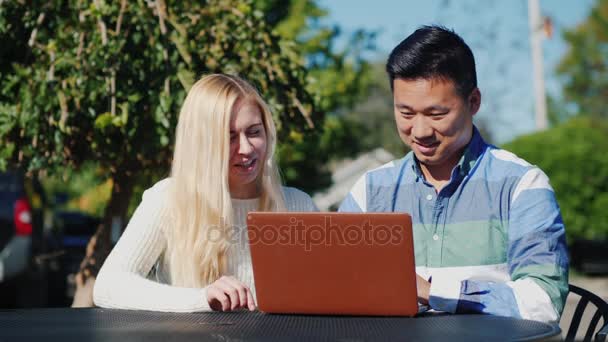 Young people caucasian woman and Chinese man are using laptop together. Have a rest on a outdoors cafe
