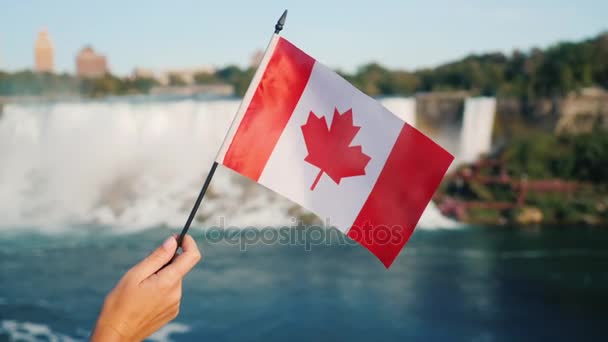 Una mano femenina sostiene una bandera canadiense en el fondo de las Cataratas del Niágara. Turismo en Canadá — Vídeos de Stock