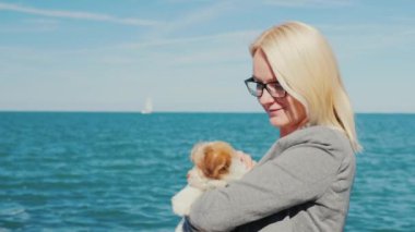 Bir kadın bir köpek yavrusu kucağında deniz arka planı tutuyor. Yatlar uzaktan görülebilir