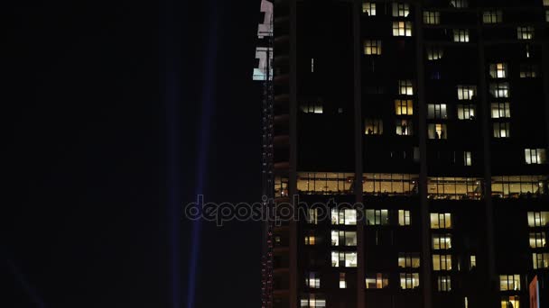 En multi-storey bostads-byggnad i mörkret. Fönstren är tända, silhuetter av människor är synliga. I den mörka himlen, en stråle av en strålkastare — Stockvideo