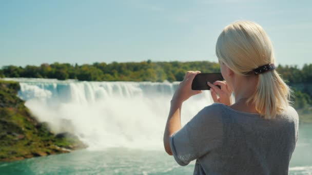 Urlaub in Kanada. Eine Touristin fotografiert die berühmten Niagarafälle. er steht an der kanadischen Küste, von wo aus der Wasserfall gut zu sehen ist — Stockvideo