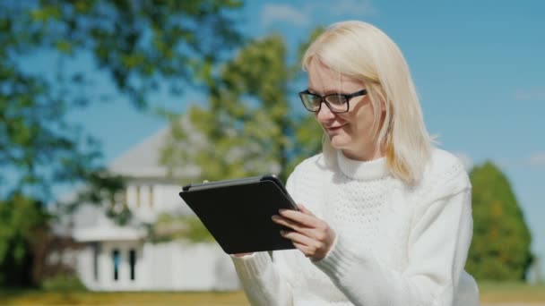 Młoda kobieta korzysta z tabletu. Siedzi na tle jego domu. Typowy podmiejskiego domu w USA — Wideo stockowe