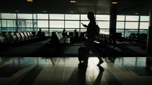 一个女人正沿着机场的终点站走着, 背着一个包在车轮上。在手智能手机 — 图库视频影像