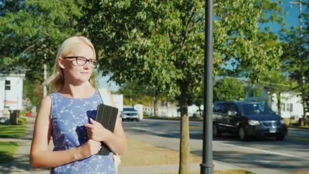 Una donna cammina tranquillamente lungo la strada di una piccola città suburbana degli Stati Uniti. Nella mano porta una tavoletta. Steadicam shot — Video Stock