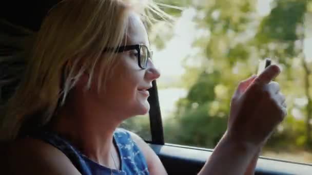 Una mujer está conduciendo en el coche en el asiento de pasajeros. Fotografía el paisaje fuera de la ventana con un smartphone — Vídeos de Stock