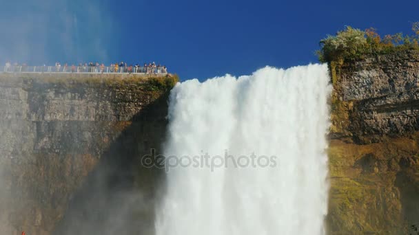 Un grupo de turistas admira las poco iluminadas Cataratas del Niágara. El aspecto inferior de disparar. En el fondo del cielo azul. Vídeo de 4K 10 bits — Vídeo de stock