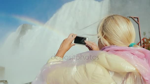 Una mujer con un impermeable está fotografiando las Cataratas del Niágara. El turismo en los EE.UU. y Canadá concepto — Vídeos de Stock