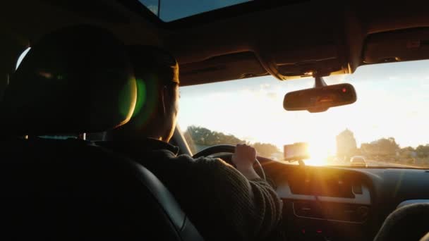 Азиат водит машину вдоль шоссе, закат солнца светит в лобовое стекло. Вид сзади — стоковое видео
