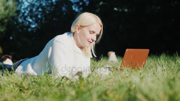 Kobieta leży na trawniku, za pomocą laptopa. Dalej biegają małe szczeniaki — Wideo stockowe