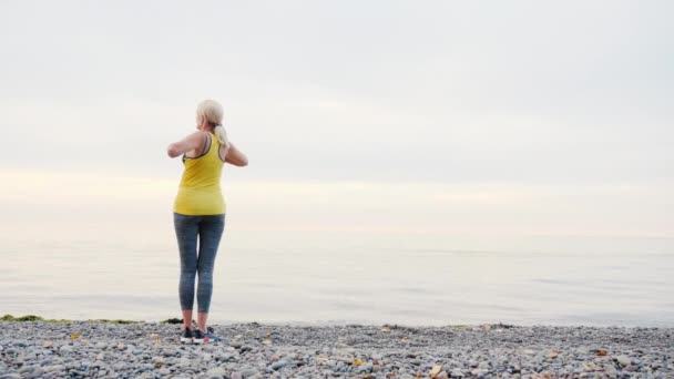 En kvinna i en gul T-shirt värms upp på stranden av en sjö eller havet — Stockvideo